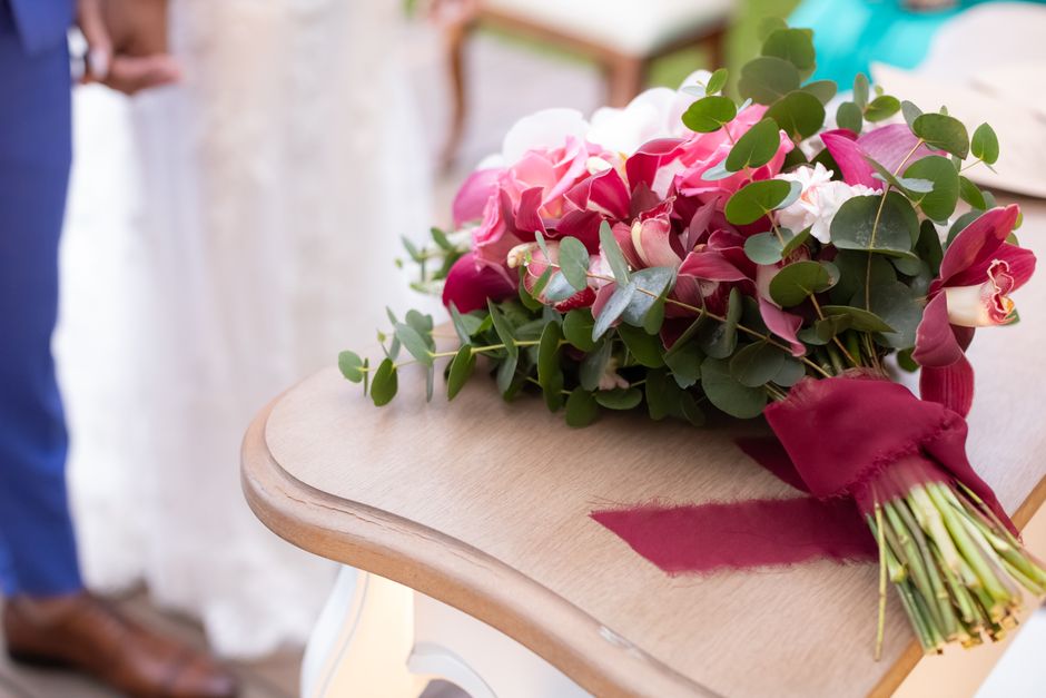 A freshly arranged floral bouquet featuring pink and white roses, complemented by sprigs of eucalyptus with round, green leaves, resting on a light wooden table. The bouquet is wrapped in deep burgund