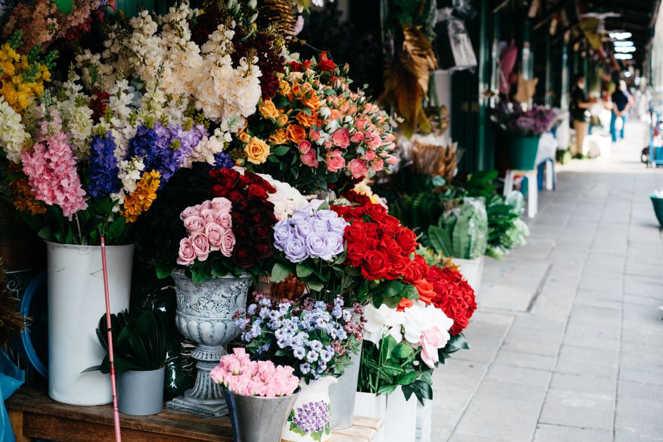 A vibrant display of fresh flowers arranged in various containers along a shopfront, featuring roses in shades of pink, red, and lavender, alongside purple and white flowers, all with lush green folia