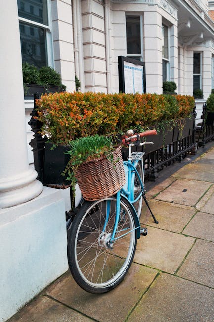 A light blue bicycle with a wicker basket attached to the front handlebar, filled with green plants and small white flowers, is parked against a black railing on a city sidewalk. Behind the bicycle, t