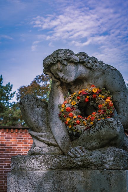 A weathered stone statue of a contemplative figure with curly hair, resting its chin on one hand and holding a circular floral wreath. The wreath is vibrant, featuring bright orange, yellow, and red f
