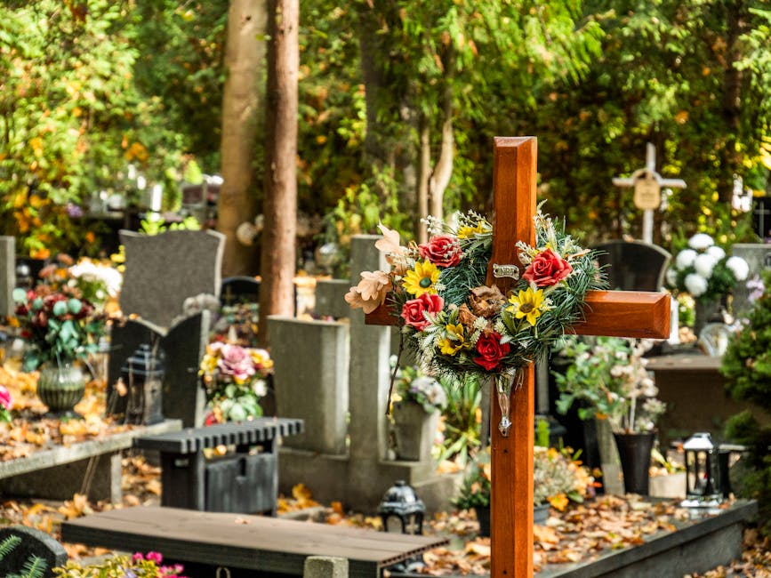 A wooden cross adorned with a floral arrangement featuring red roses, yellow daisies, beige lilies, and green foliage, placed among rows of memorial flowers and wreaths in a cemetery. The background i