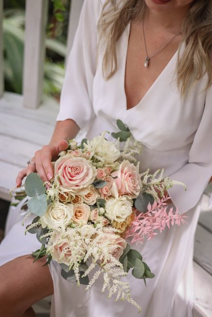 A woman dressed in a white V-neck dress holds a delicate floral bouquet consisting of pastel pink and cream roses, complemented by white and light green filler flowers, and soft eucalyptus leaves. The