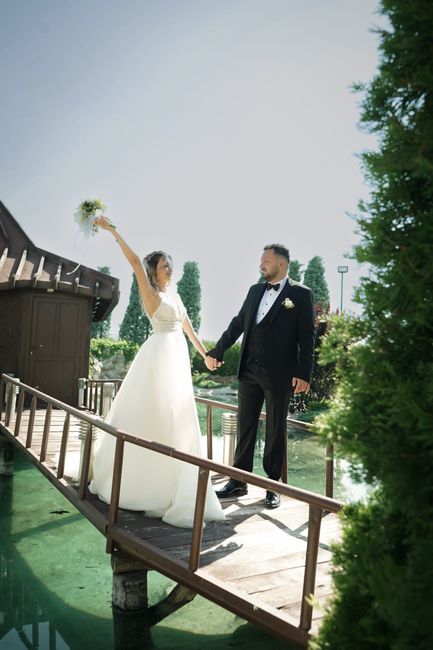 A bride and groom stand on a small wooden bridge outdoors, holding hands. The bride wears a white wedding gown with lace details and holds a bouquet of white and green flowers raised in the air. The g