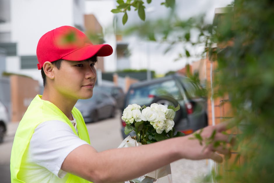 A young delivery person wearing a red cap and a high-visibility yellow vest is placing a bouquet of white hydrangeas wrapped in white paper into a bush or shrubbery outside a residential building. The