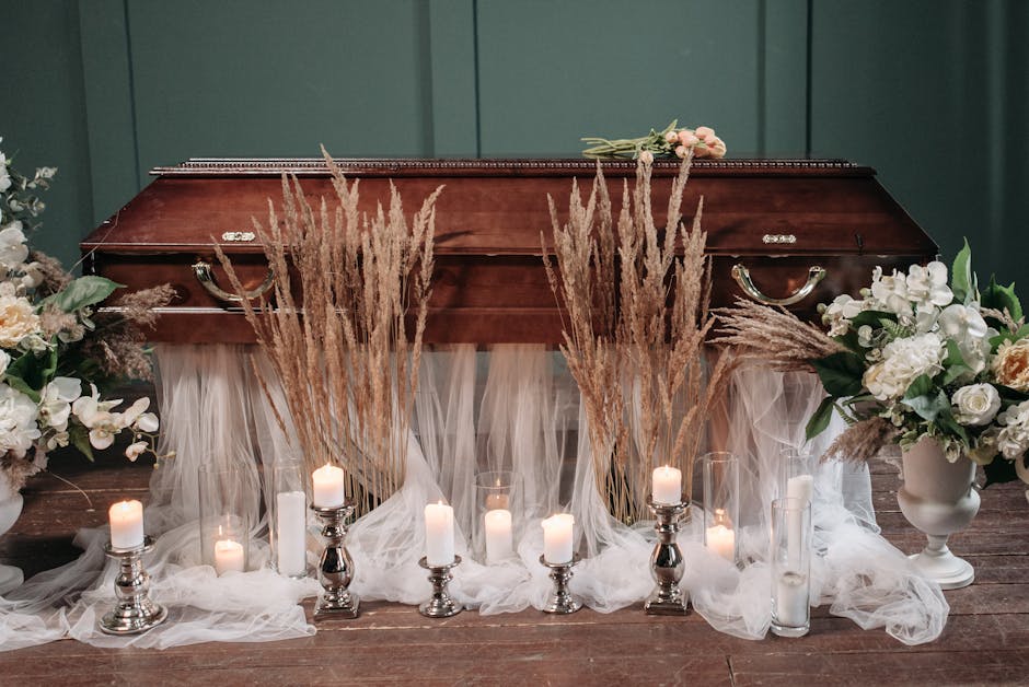 A wooden casket surrounded by floral arrangements featuring white roses, lilies, and greenery, set against a dark green wall. Tall, dried pampas grass stems are placed in front of the casket, with sof