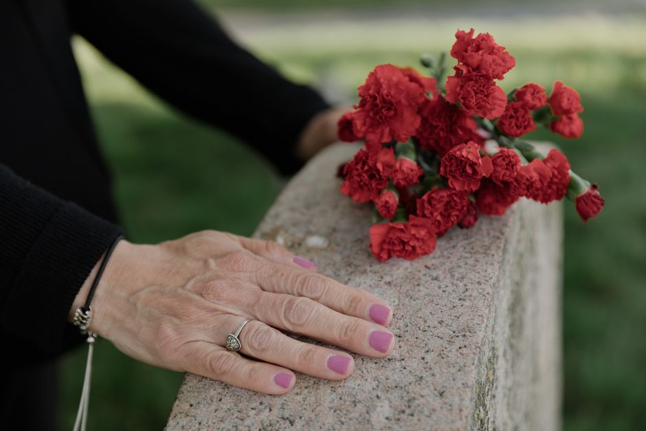 A close-up of a person's hand resting on a stone ledge, showing pink-painted nails and a silver ring with a large, sparkling stone. Next to the hand, on the stone surface, is a bouquet of red carnatio