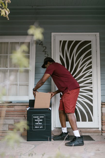 A delivery person wearing a red T-shirt, red shorts, black boots, and a cap is placing a floral gift into a black parcel drop box outside a residential building with light blue wooden siding, a white-