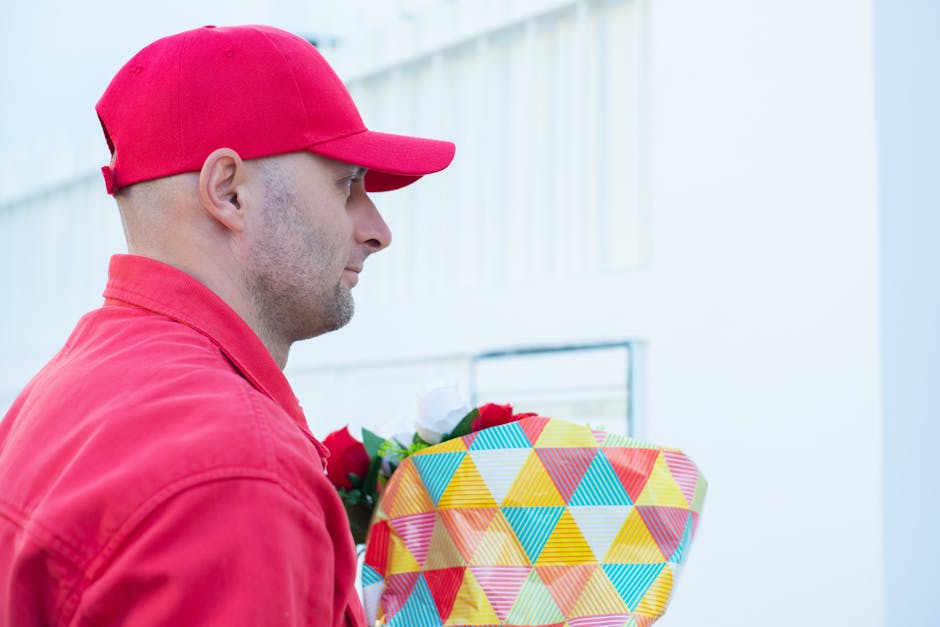 A delivery person wearing a red cap and red jacket is holding a vibrant floral gift wrapped in multicolored geometric patterned paper with a yellow ribbon. The bouquet features a mix of fresh flowers,
