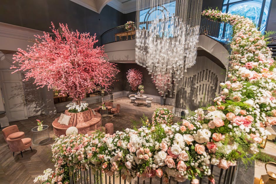 Elegant interior decorated for a wedding event with large pink cherry blossom trees and cascading floral arrangements of pink and white roses, peonies, and hydrangeas. The arrangement surrounds a cent