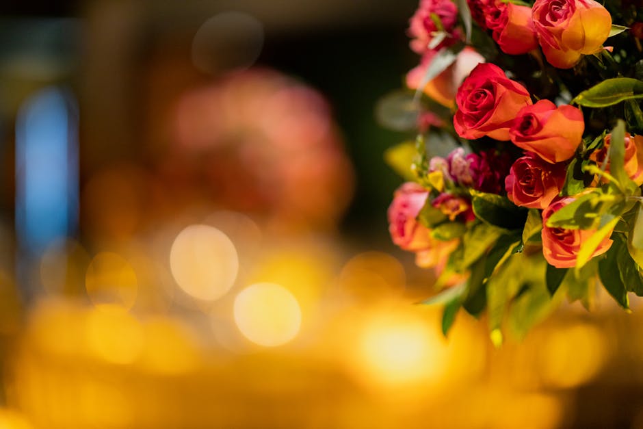 A close-up view of a bouquet of fresh roses in shades of pink, orange, and yellow, arranged with lush green leaves. The flowers are beautifully clustered, with some petals slightly curled, indicating